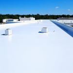 White commercial flat roof with rooftop vents under a bright blue sky, surrounded by trees and nearby industrial buildings.