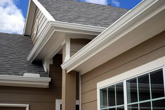 White gutters along a tan home's roofline with gray shingles, clean trim, and a window beneath a partly cloudy sky.