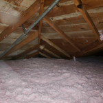 Attic filled with pink blown-in insulation beneath wood roof trusses, showing roof decking and utility piping overhead.