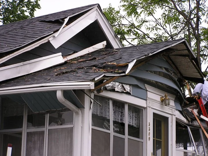 Storm-damaged home with torn shingles, bent gutters, exposed roof edges, and a worker on a ladder near the damaged entry.