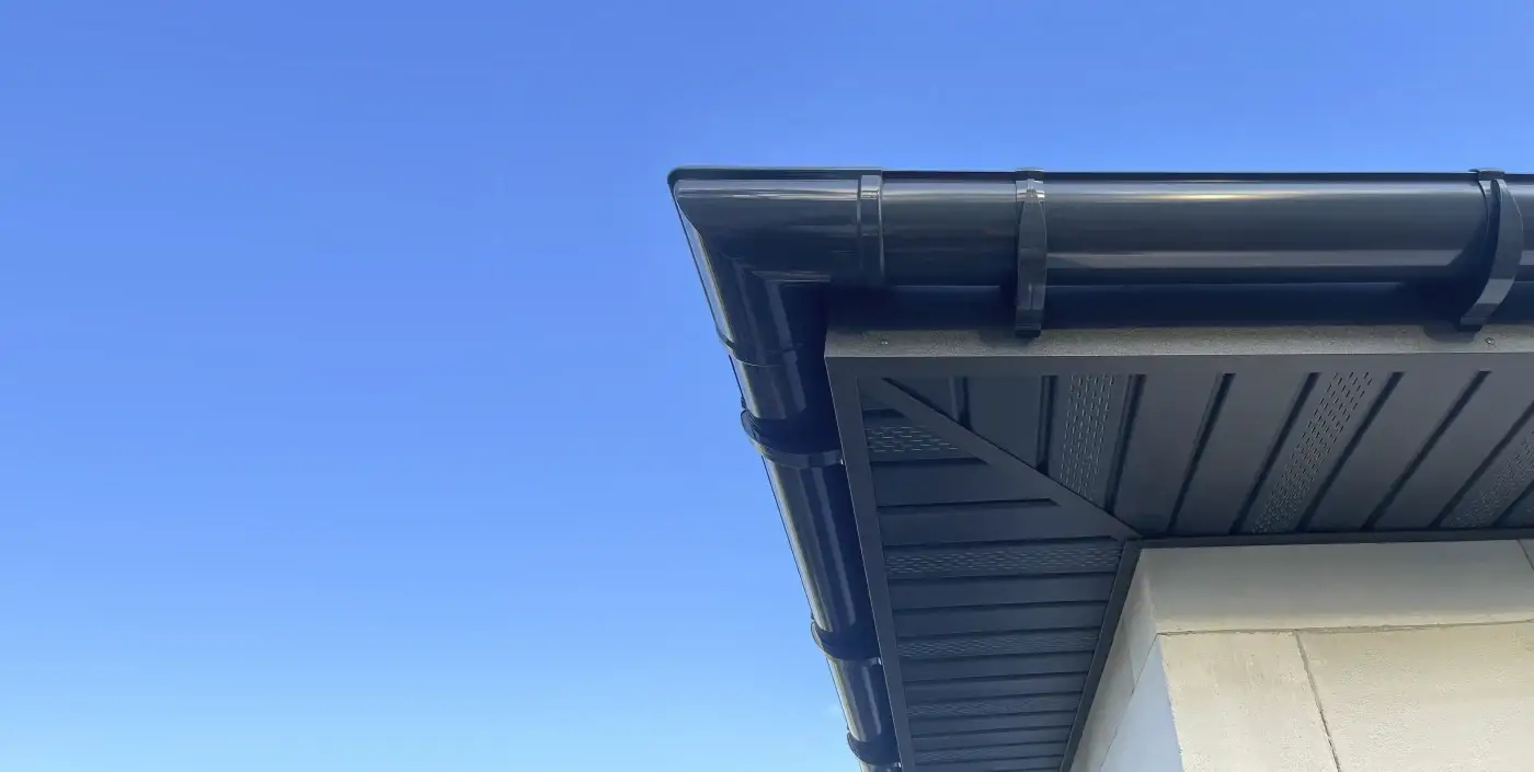 Black commercial gutter and downspout installed under a roof edge with vented soffit, pale wall, and clear blue sky above.